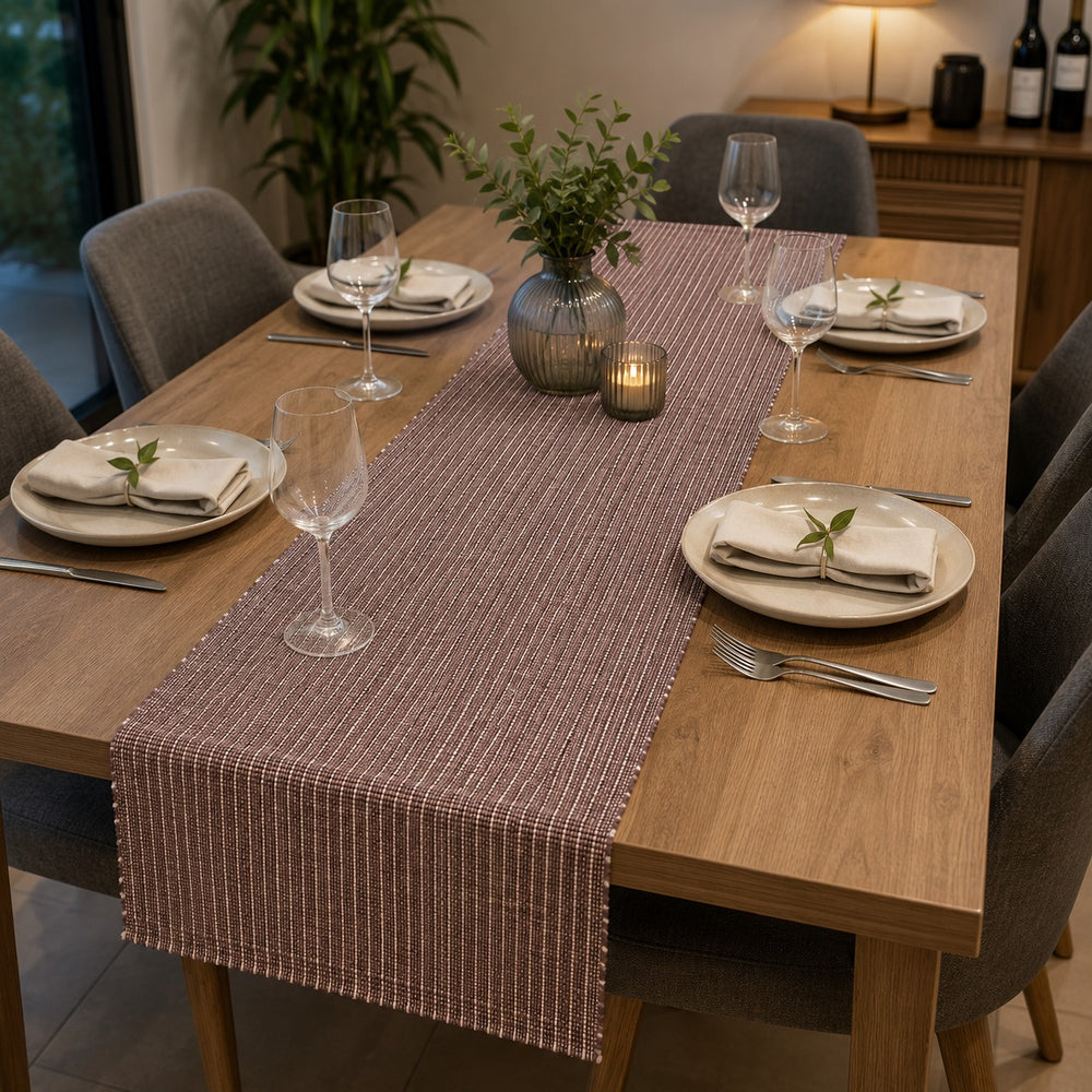 A rectangular woven table runner in a muted mauve-brown color with thin vertical white stripes, styled on a wooden dining table with plates, wine glasses, and a small vase of greenery.