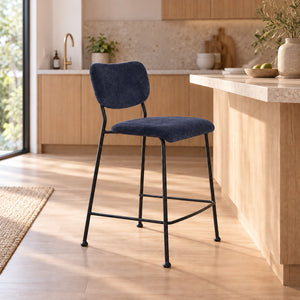 A modern bar stool with a navy blue velvet upholstered seat and backrest, supported by a black metal frame, set in a bright, contemporary kitchen.