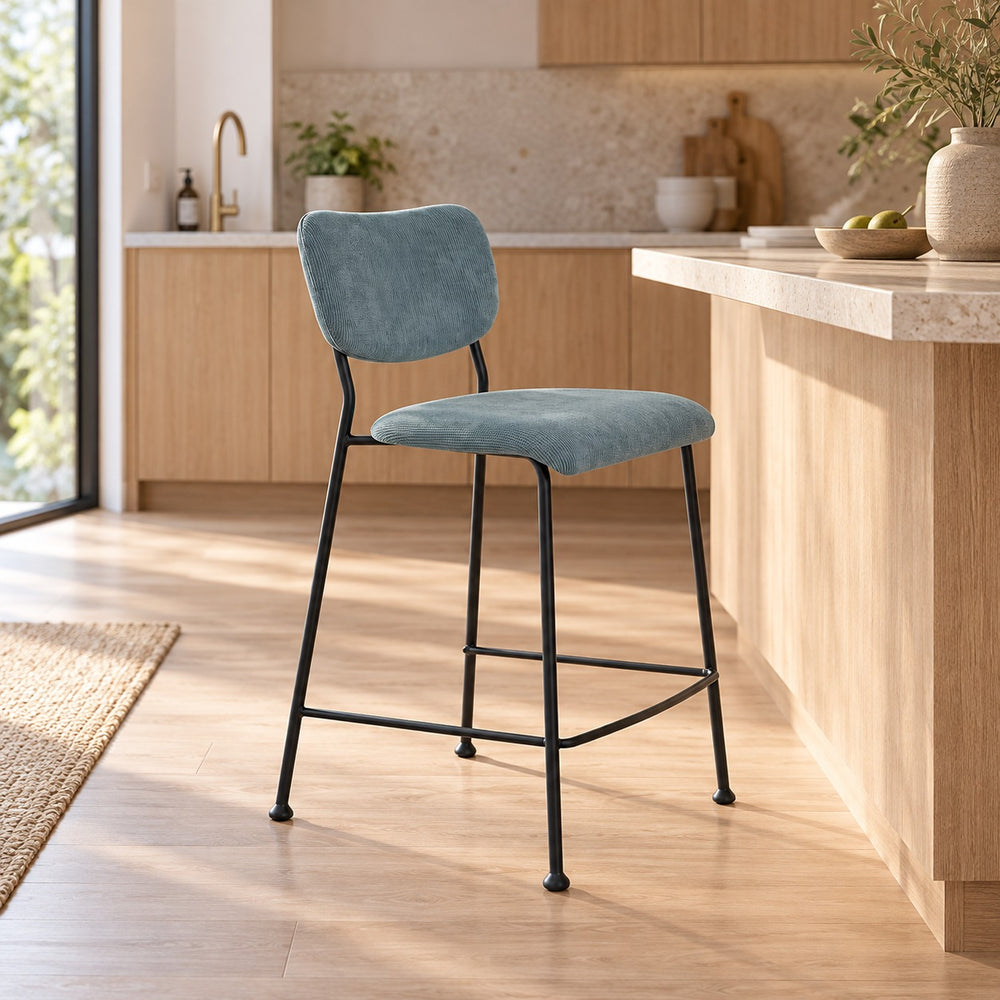 A modern bar stool with a dusty pink velvet upholstered seat and backrest, supported by a slim black metal frame, set in a bright, contemporary kitchen.