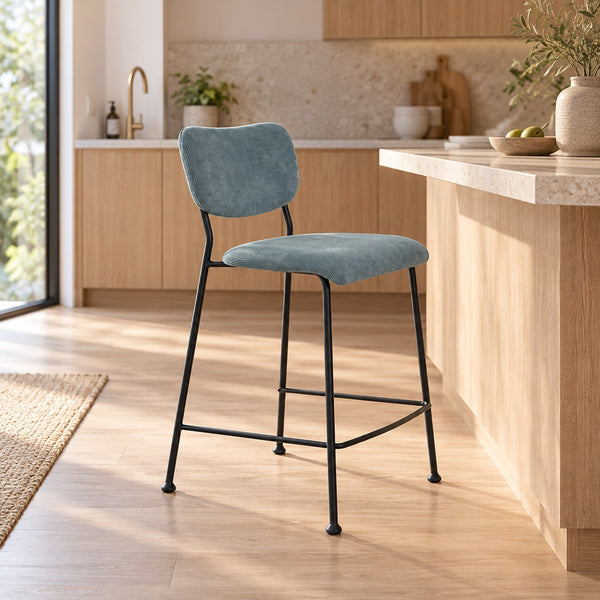 A modern bar stool with a light blue velvet upholstered seat and backrest, supported by a slim black metal frame, positioned in a bright, contemporary kitchen setting.