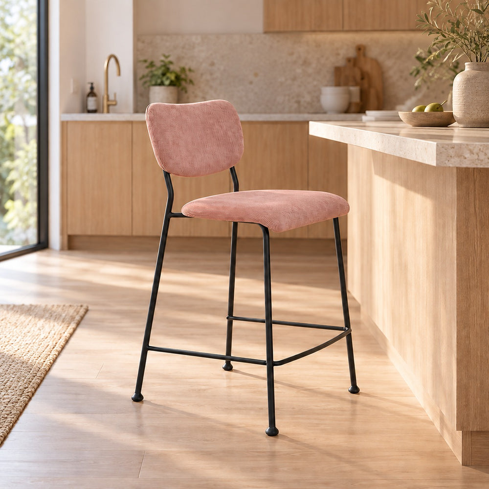 A modern bar stool with a dusty pink velvet upholstered seat and backrest, supported by a slim black metal frame, set in a bright, contemporary kitchen.