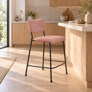 A modern bar stool with a dusty pink velvet upholstered seat and backrest, supported by a slim black metal frame, set in a bright, contemporary kitchen.