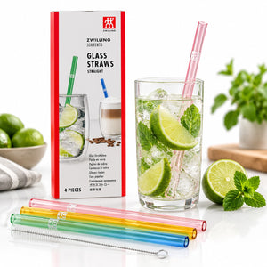 Four colorful glass drinking straws with a metal cleaning tool, shown alongside their white and red packaging box and a glass of iced limeade with mint and a pink straw. The scene is styled with limes, mint leaves, and a potted plant on a white background.