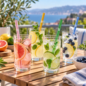 Four tall clear drinking glasses filled with iced cocktails and garnishes, arranged on a wooden table outdoors with a beach and ocean background. The glasses have colorful straws and contain pink, yellow, mint, and blue-toned drinks with citrus and berry garnishes.