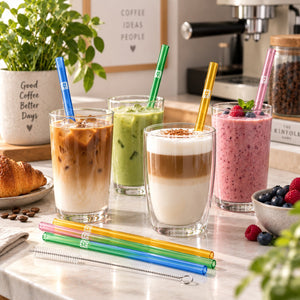 Four colorful reusable glass straws in blue, green, yellow, and pink are shown with four different iced and layered drinks on a kitchen counter, with coffee-themed decor in the background.