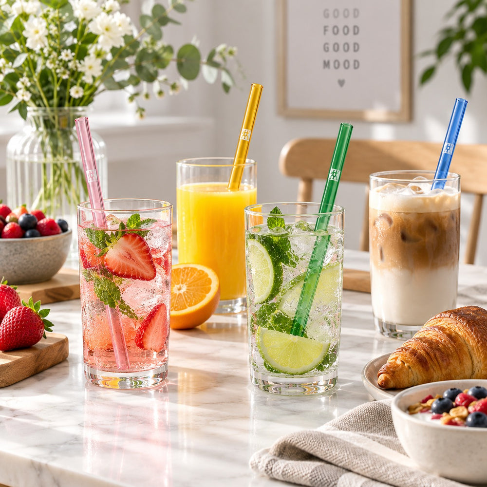 Four colored glass straws in pink, yellow, green, and blue are shown in a clear drinking glass with lime slices and mint, alongside their product packaging box and a metal straw cleaning tool on a marble surface.